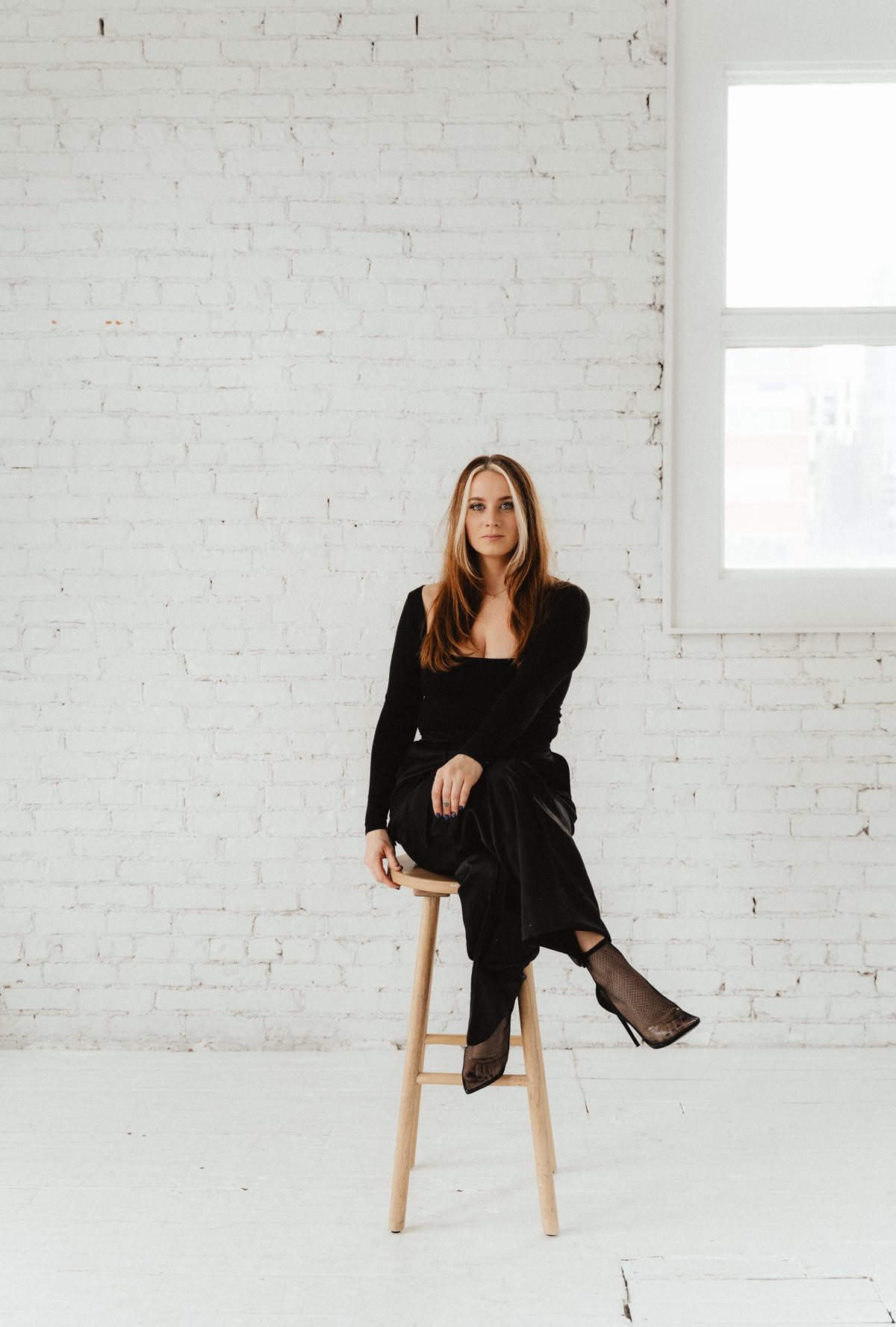 Becky Pedersen seated on a wooden stool in front of a white brick wall